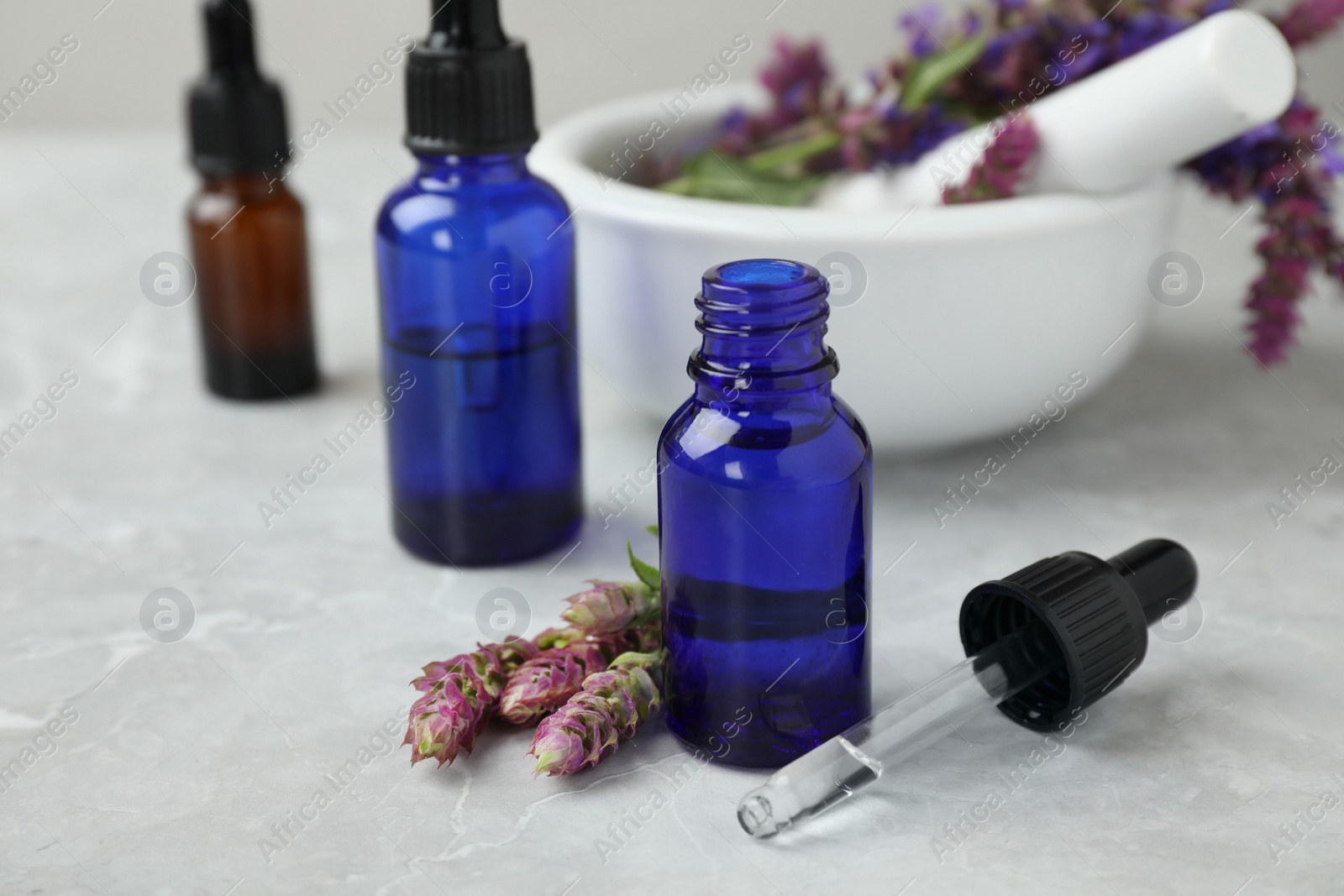 Photo of Bottles of sage essential oil and flowers on grey table