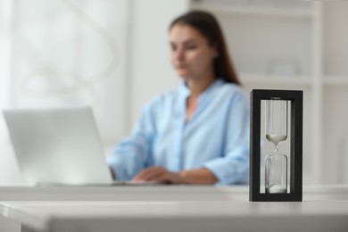 Hourglass with flowing sand on white table. Woman using laptop indoors, selective focus