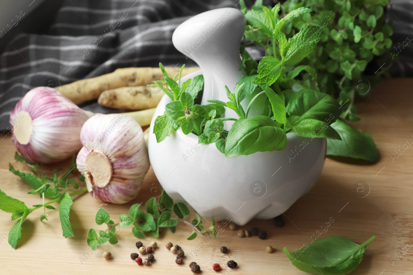 Photo of Mortar with different fresh herbs near garlic, horseradish roots and black peppercorns on wooden table, closeup