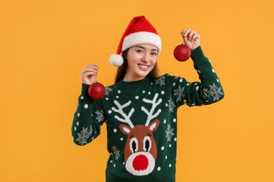Happy young woman in Christmas sweater and Santa hat holding festive baubles on orange background
