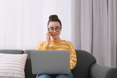 Home workplace. Happy woman with laptop talking on smartphone on sofa in room