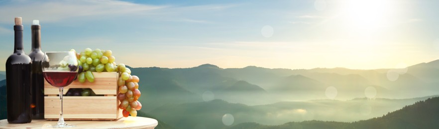 Tasty wine and grapes on wooden table against beautiful mountain landscape, space for text. Banner design