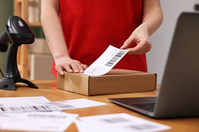 Photo of Parcel packing. Post office worker sticking barcode on box at wooden table indoors, closeup