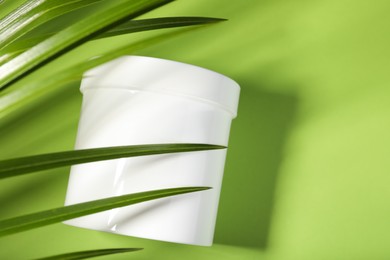 Blank jar of body cream with palm leaf on light green background, top view