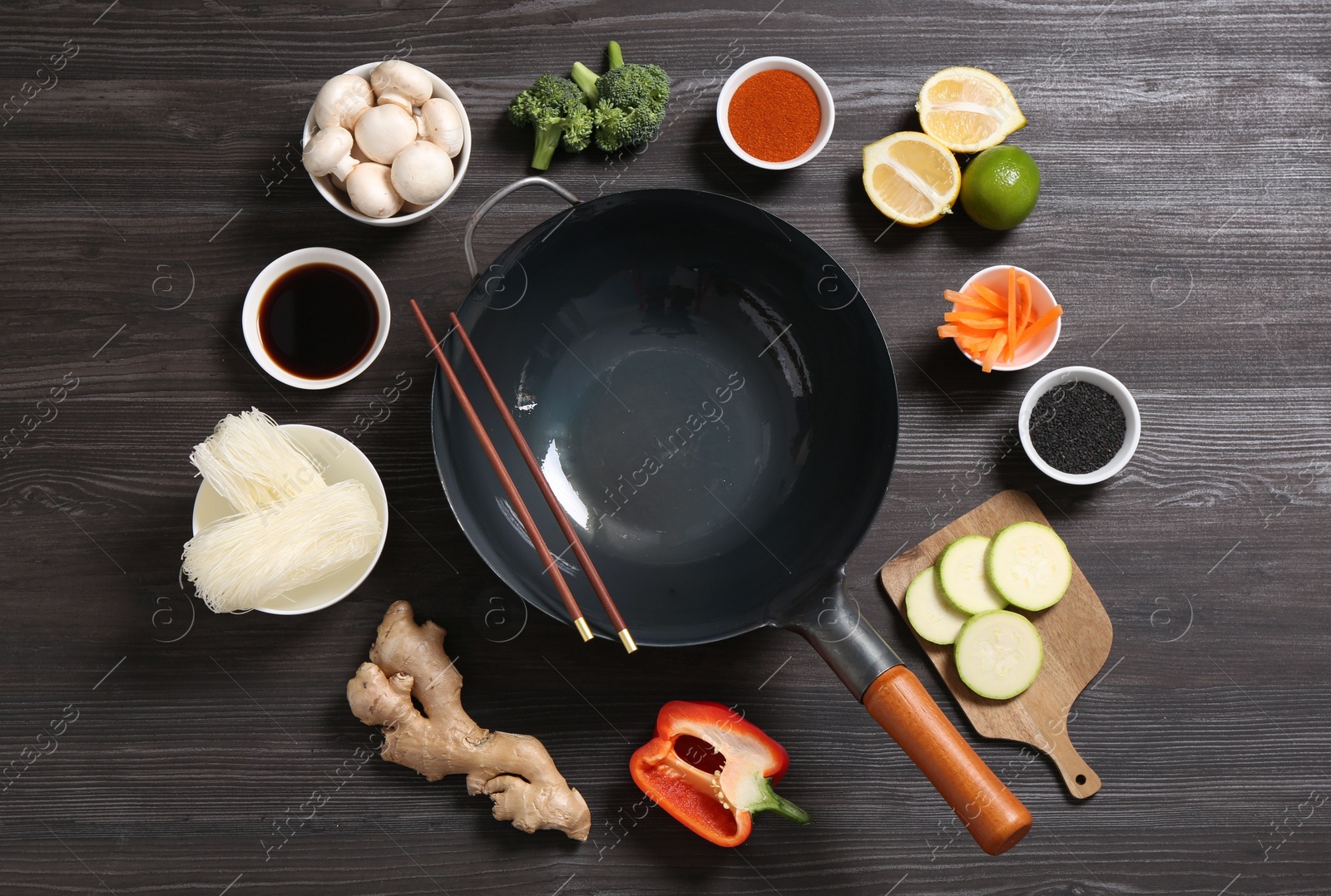 Photo of Empty iron wok and chopsticks surrounded by ingredients on dark grey wooden table, flat lay