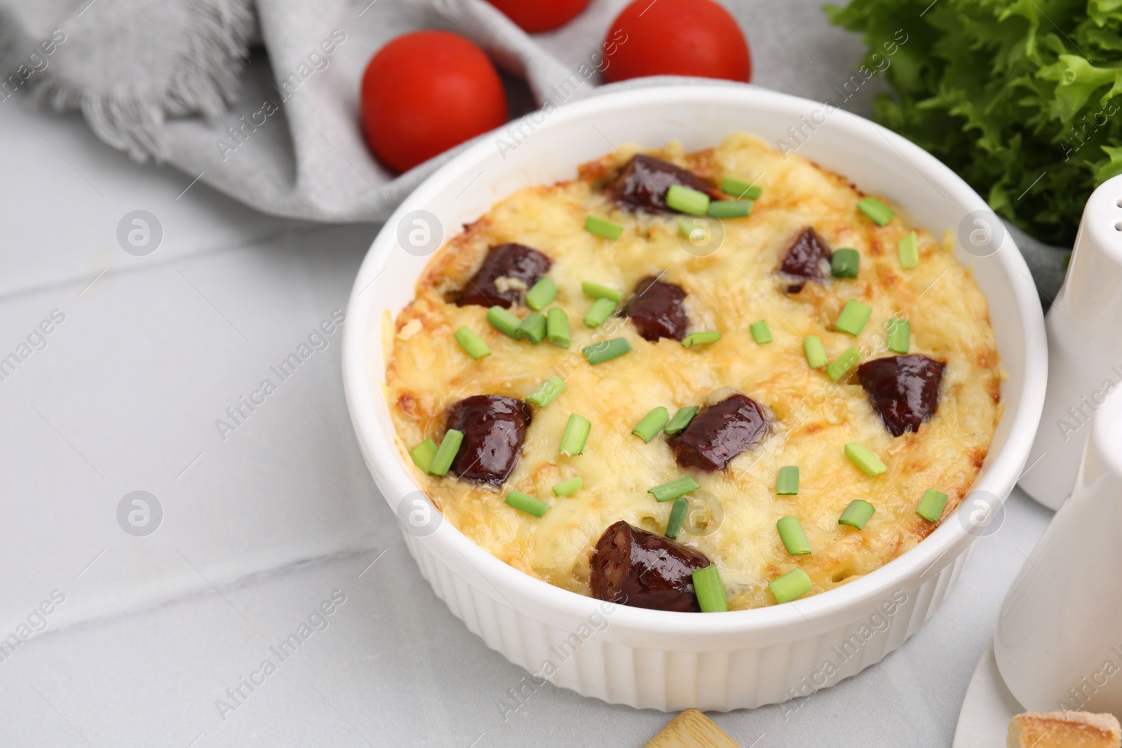 Photo of Tasty sausage casserole with green onions in baking dish served on white tiled table, closeup