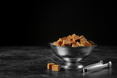 Photo of Metal bowl with brown sugar cubes and tongs on black table. Space for text
