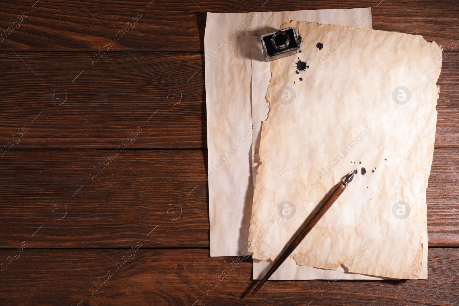 Photo of Fountain pen, inkwell and vintage parchment with ink stains on wooden table, flat lay. Space for text