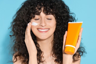 Photo of Beautiful young woman with tube of sunscreen applying sun protection cream onto her face on light blue background