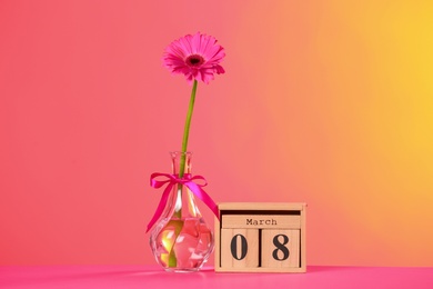 Photo of Vase with beautiful flower and wooden block calendar on table against color background. International Women's Day