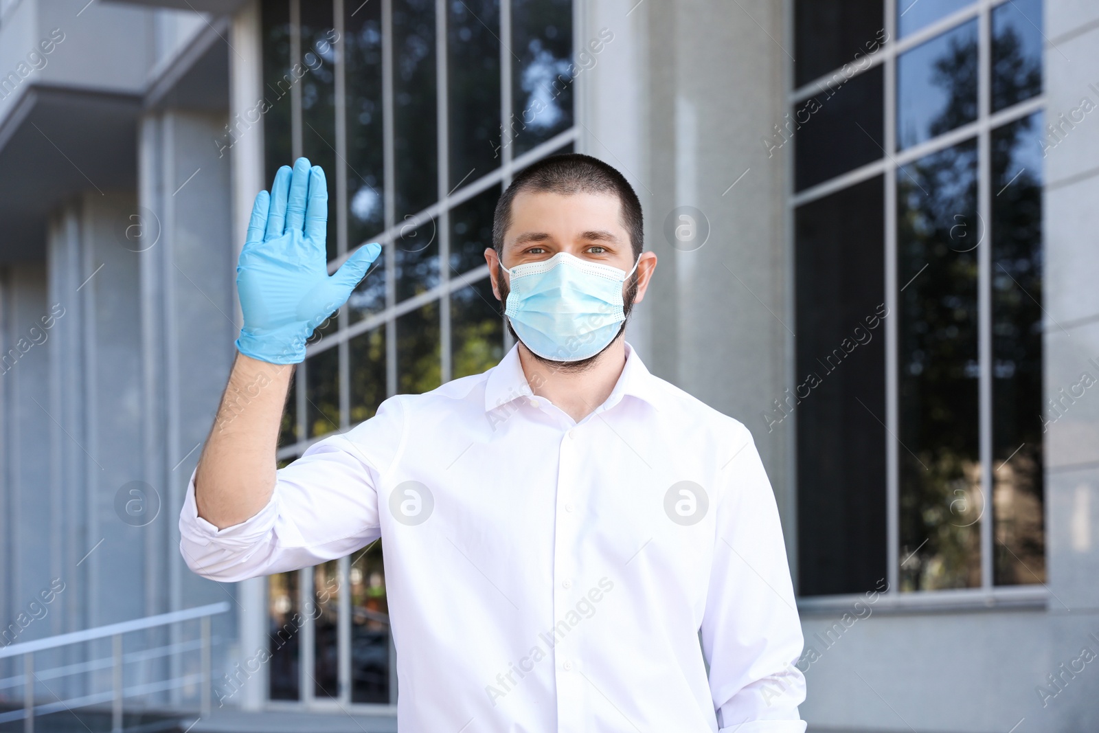 Photo of Man in protective face mask showing hello gesture outdoors. Keeping social distance during coronavirus pandemic