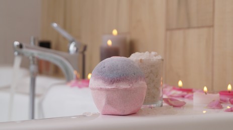 Colorful bath bomb, sea salt, flower petals and burning candles on white tub in bathroom