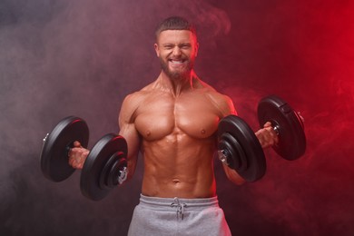 Photo of Emotional young bodybuilder exercising with dumbbells in smoke on color background