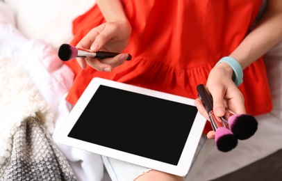 Photo of Young woman with makeup brushes using tablet on bed. Beauty blogger