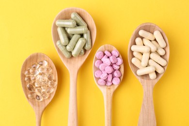 Different vitamin pills in wooden spoons on yellow background, flat lay