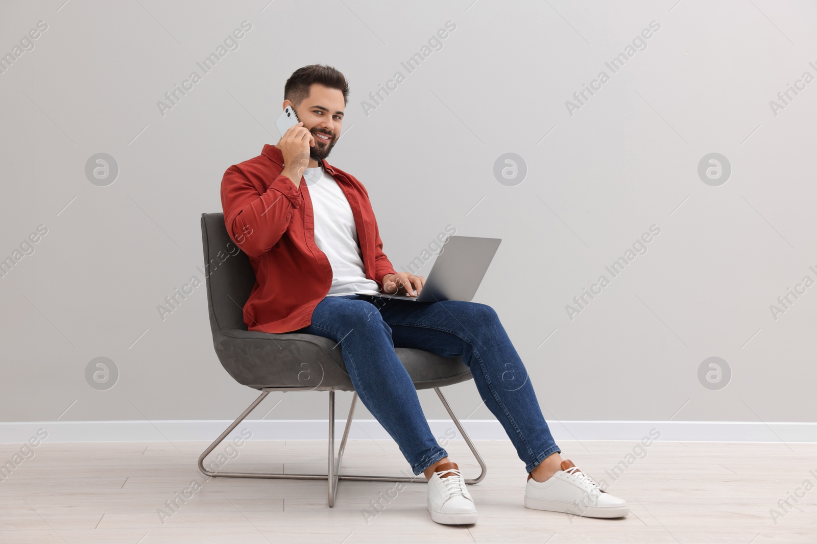 Photo of Handsome man with laptop talking on smartphone while sitting in armchair near light grey wall indoors