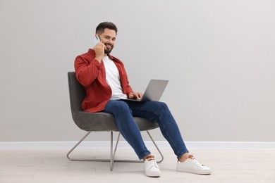 Photo of Handsome man with laptop talking on smartphone while sitting in armchair near light grey wall indoors