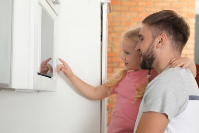Photo of Little daughter and father using microwave oven in kitchen