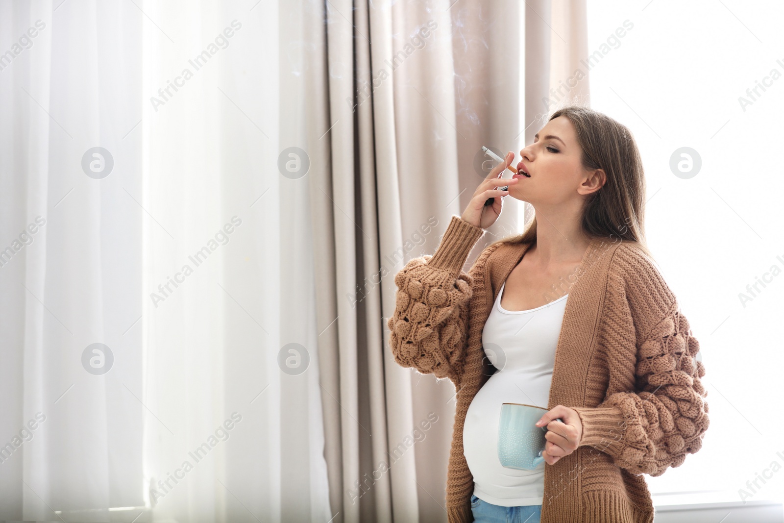 Photo of Young pregnant woman with cup of coffee smoking cigarette at home. Harm to unborn baby