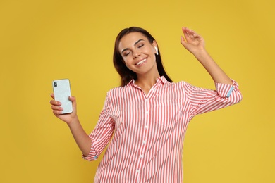 Happy young woman with smartphone listening to music through wireless earphones on yellow background