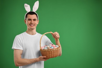 Photo of Easter celebration. Handsome young man with bunny ears holding basket of painted eggs on green background. Space for text
