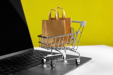 Photo of Online store. Laptop, mini shopping cart and purchases on beige table, closeup