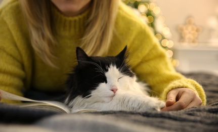 Photo of Woman reading book and hugging adorable cat on grey blanket, closeup