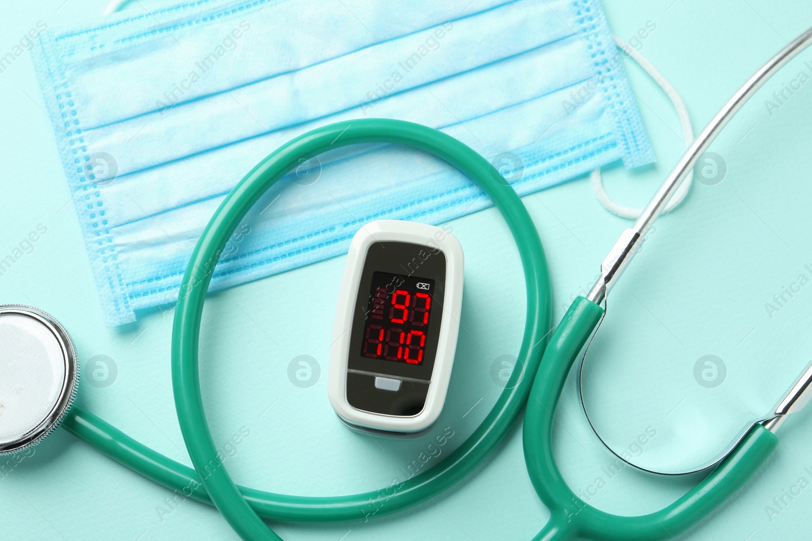 Photo of Modern fingertip pulse oximeter, mask and stethoscope on light blue background, flat lay