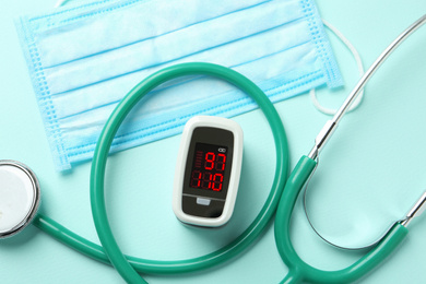 Photo of Modern fingertip pulse oximeter, mask and stethoscope on light blue background, flat lay