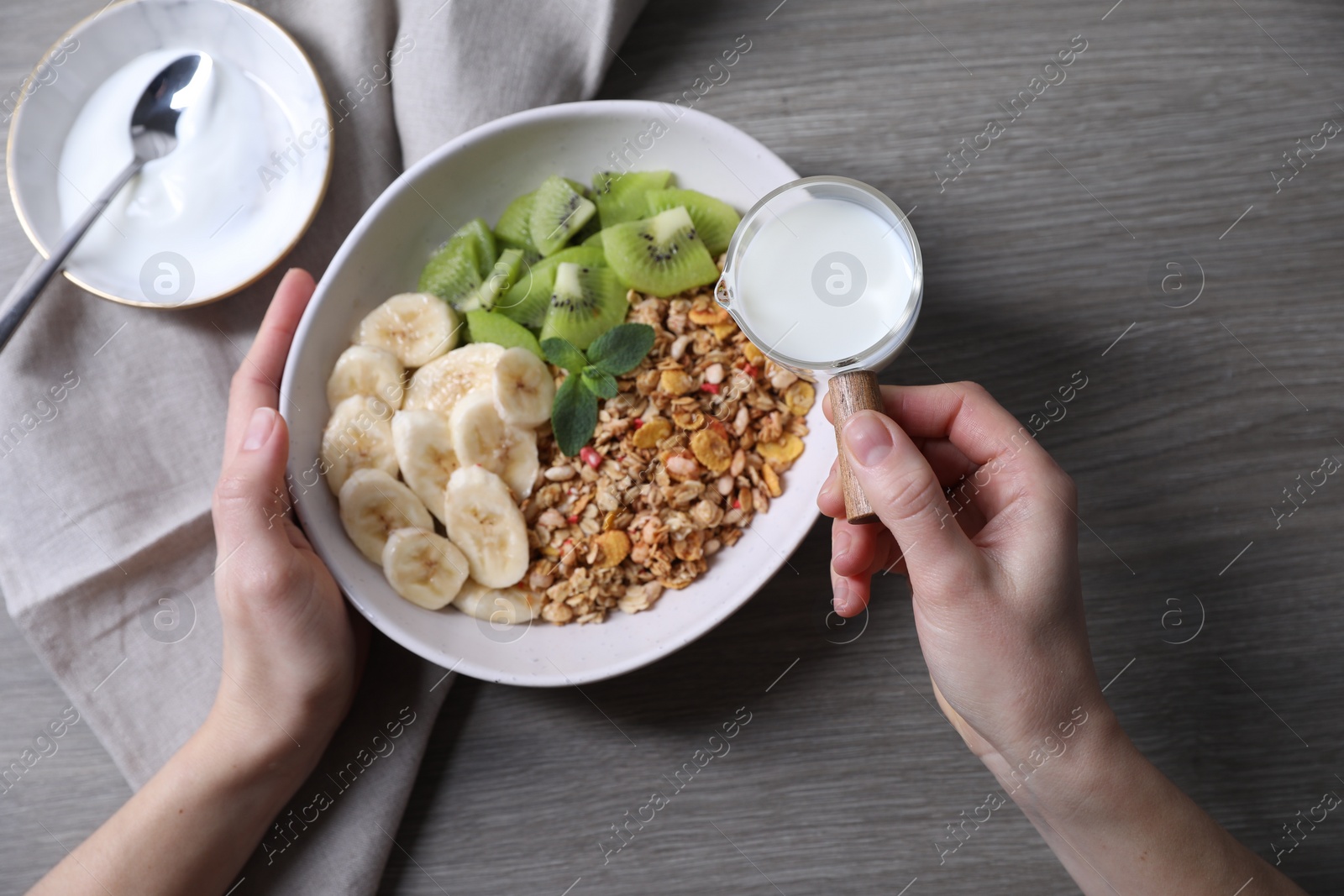 Photo of Woman pouring milk into bowl of tasty granola with banana and kiwi at grey wooden table, top view