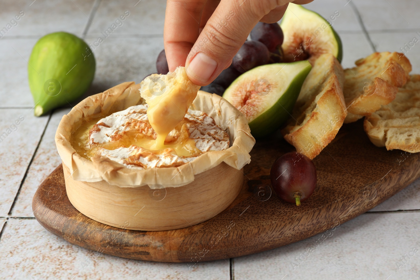 Photo of Woman dipping bread into baked brie cheese at light tiled table, closeup