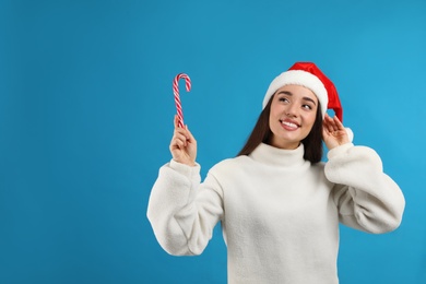 Photo of Young woman in beige sweater and Santa hat holding candy cane on light blue background, space for text. Celebrating Christmas