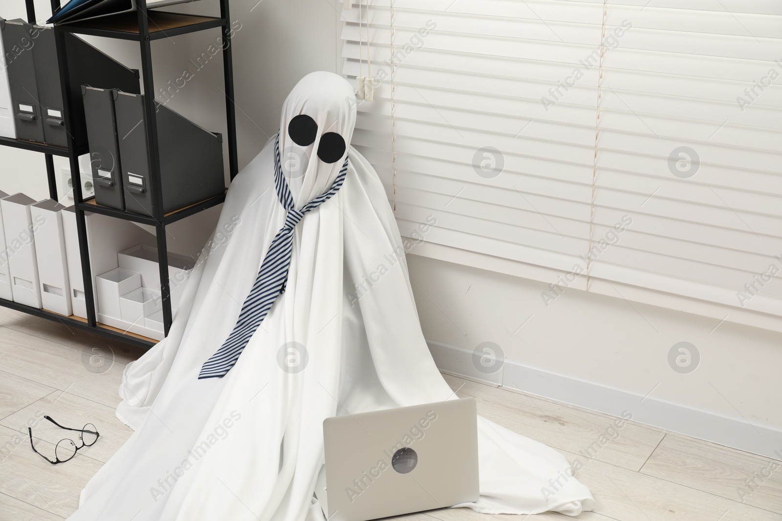 Photo of Overworked ghost. Man in white sheet with laptop on floor in office, space for text