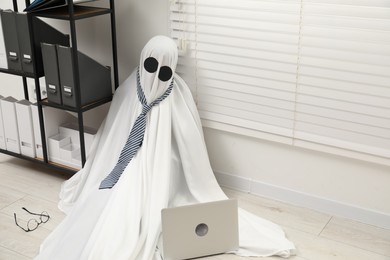 Photo of Overworked ghost. Man in white sheet with laptop on floor in office, space for text