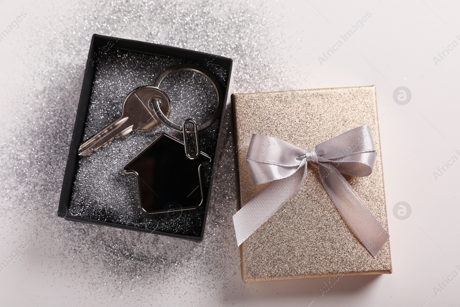 Photo of Key with trinket in shape of house, glitter and gift box on light grey background, flat lay. Housewarming party