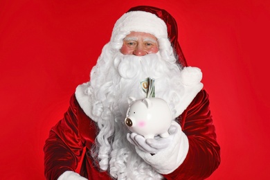 Photo of Santa Claus holding piggy bank with dollar banknotes on red background