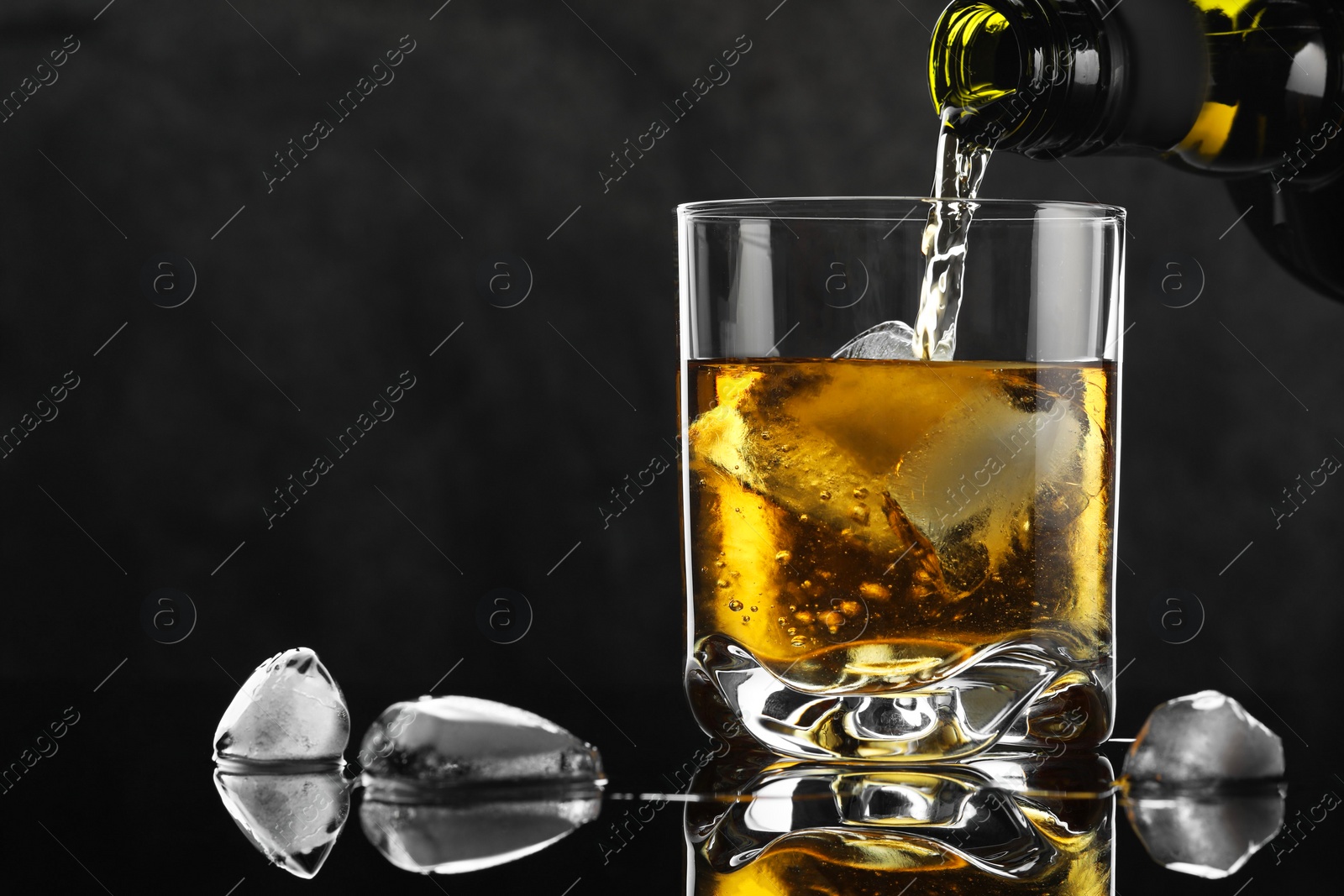 Photo of Pouring tasty whiskey from bottle into glass with ice at mirror table against black background, closeup. Space for text