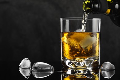 Pouring tasty whiskey from bottle into glass with ice at mirror table against black background, closeup. Space for text