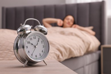 Photo of Alarm clock on table. Woman stretching in soft bed at home, selective focus