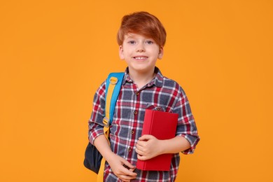 Happy schoolboy with backpack and book on orange background