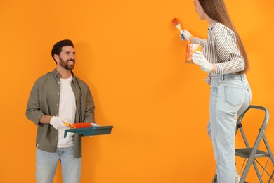 Photo of Woman painting orange wall and man holding container with roller. Interior design