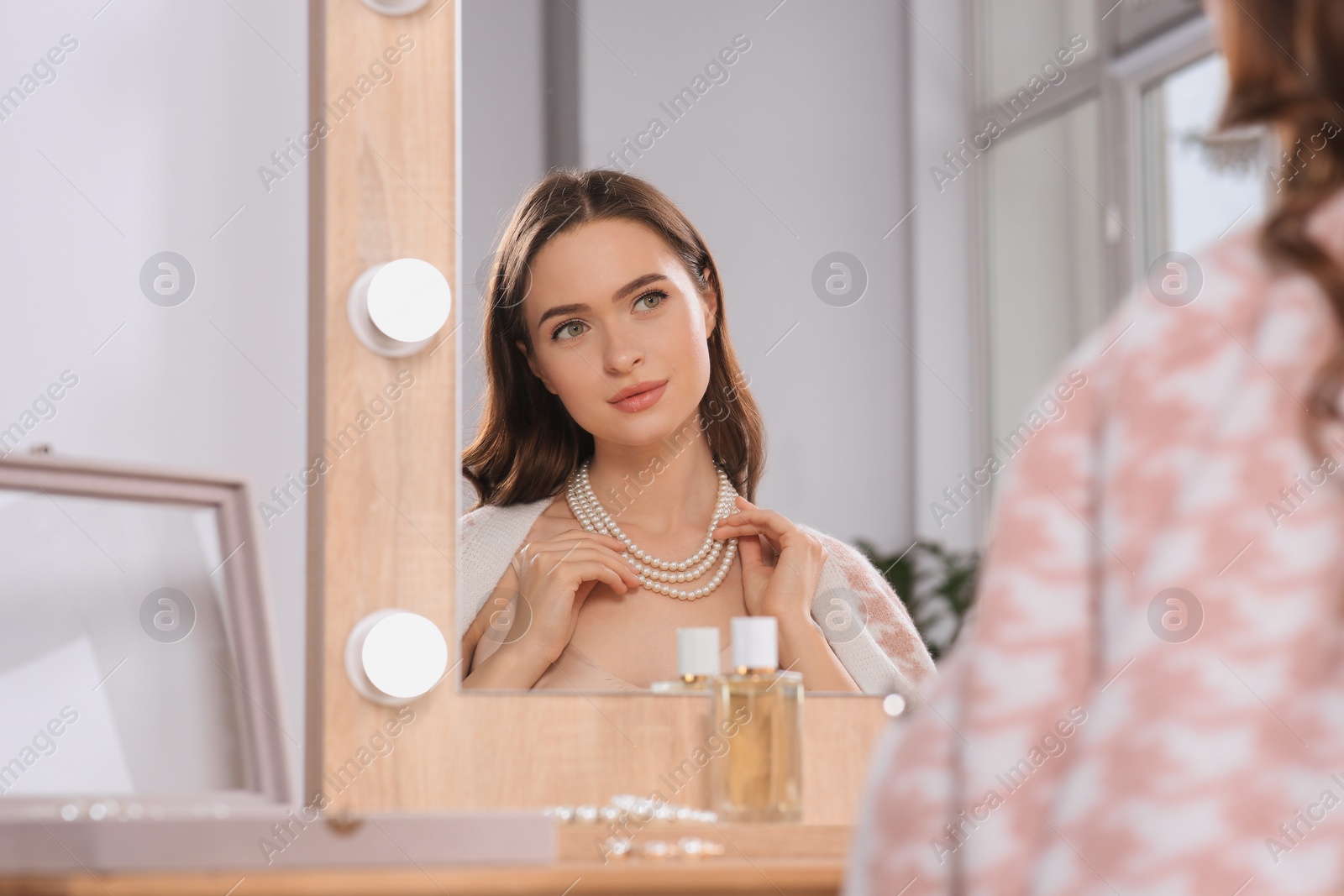 Photo of Young woman wearing elegant pearl necklace near mirror at dressing table indoors