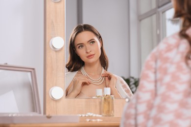 Young woman wearing elegant pearl necklace near mirror at dressing table indoors