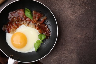 Fried egg, bacon and basil in frying pan on brown table, top view. Space for text