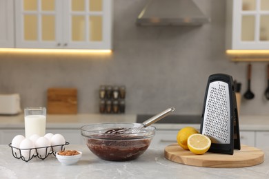 Photo of Metal whisk, chocolate cream in bowl, grater and different products on gray marble table indoors