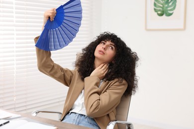 Photo of Young businesswoman waving blue hand fan to cool herself at table in office