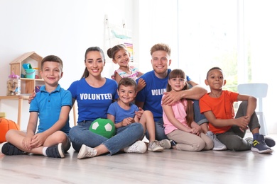 Photo of Happy volunteers with children sitting on floor indoors