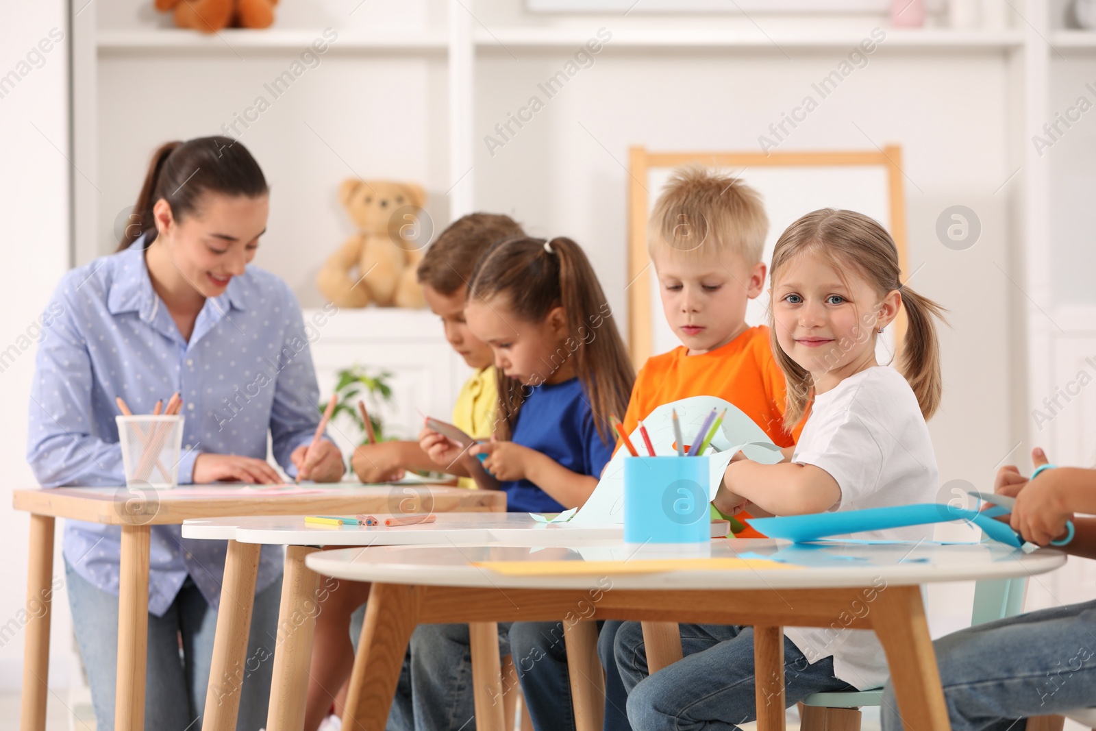 Photo of Nursery teacher with group of cute little children drawing and cutting paper at desks in kindergarten. Playtime activities