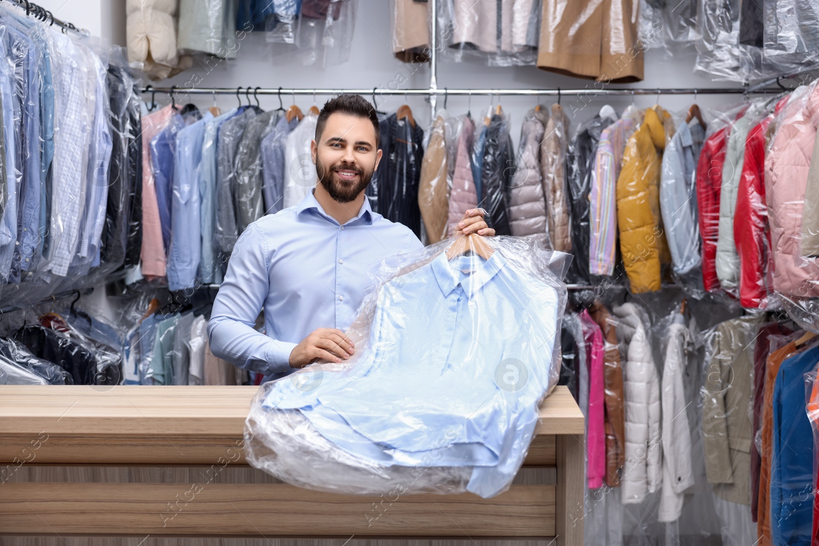 Photo of Dry-cleaning service. Happy worker holding hangers with clothes in plastic bags at counter indoors