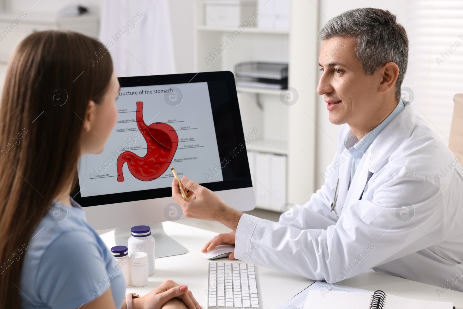 Photo of Gastroenterologist showing screen with illustration of human stomach model to patient at table in clinic